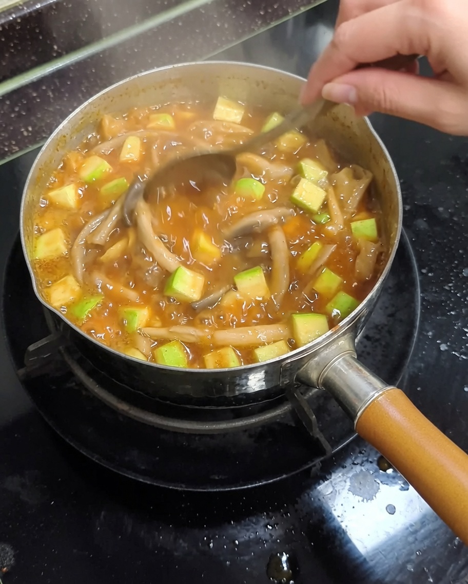 A metal pot on a stovetop with boiling soybean paste stew being stirred with a metal spoon, showing zucchini and mushrooms.