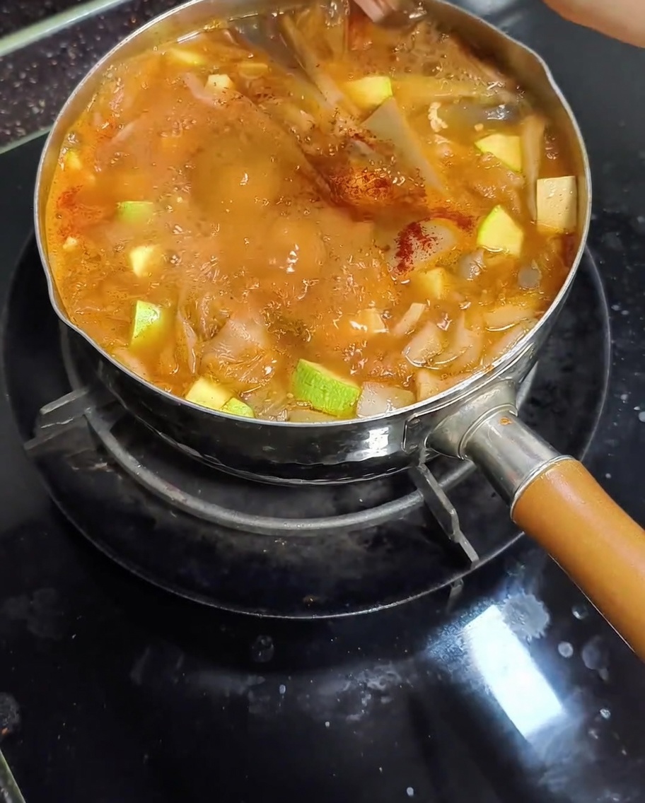 Red sweet paprika powder being sprinkled into a pot of boiling Korean soybean paste stew containing zucchini and mushrooms.