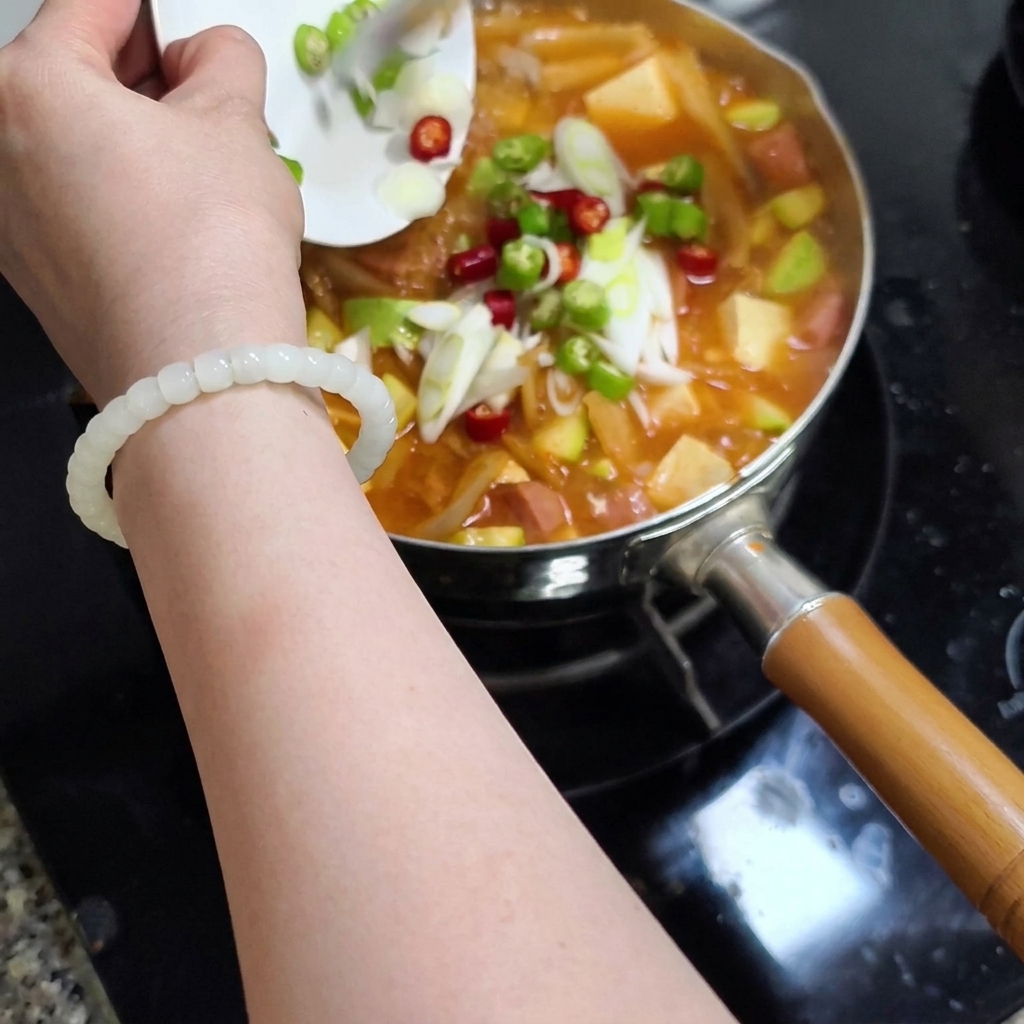 A hand holding a small white plate sliding chopped green and red chili peppers and sliced scallions into a pot of bubbling stew.
