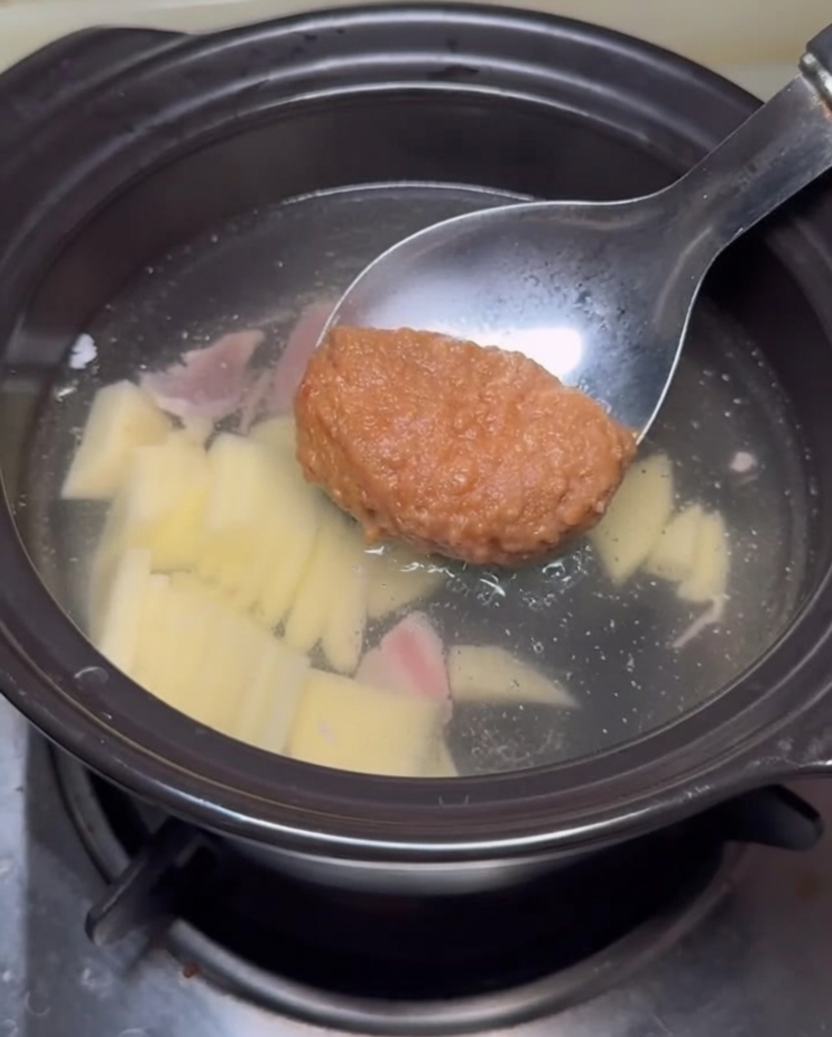 A ladle holding a dollop of thick brown soybean paste being submerged into the stew broth.