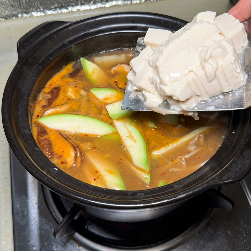 Silken tofu blocks being slid from a cleaver into the hot, bubbling soybean paste stew.