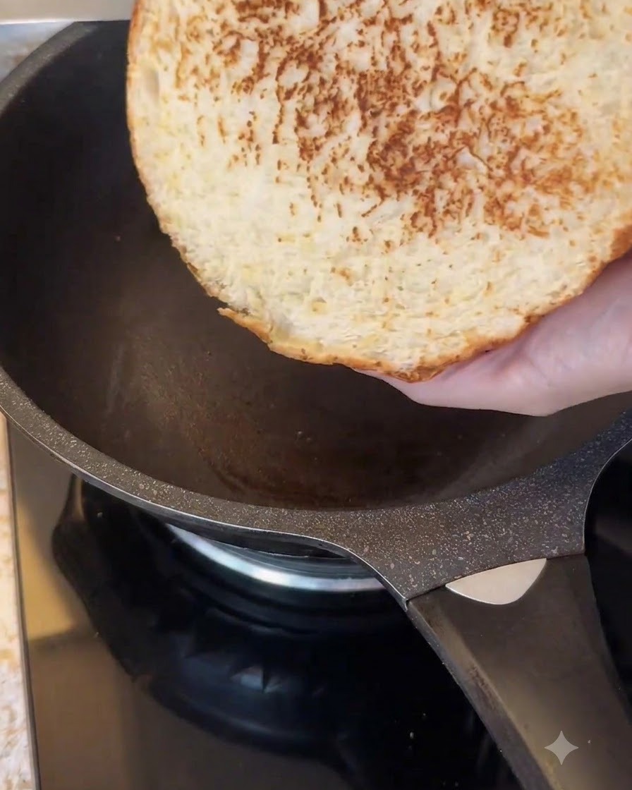 The cut side of a large burger bun being toasted in a dark non-stick frying pan.