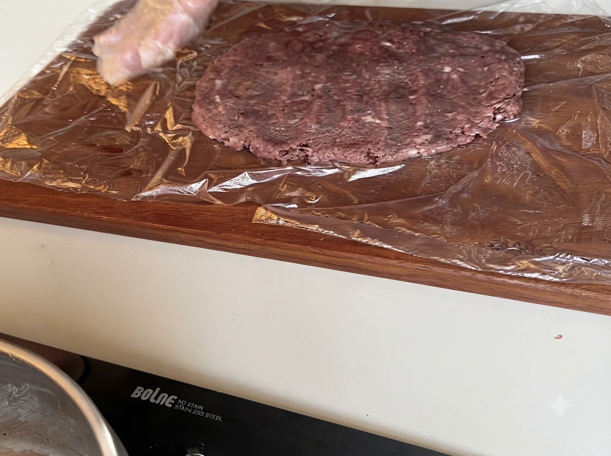 A large raw beef patty being flattened on plastic wrap spread over a wooden cutting board.