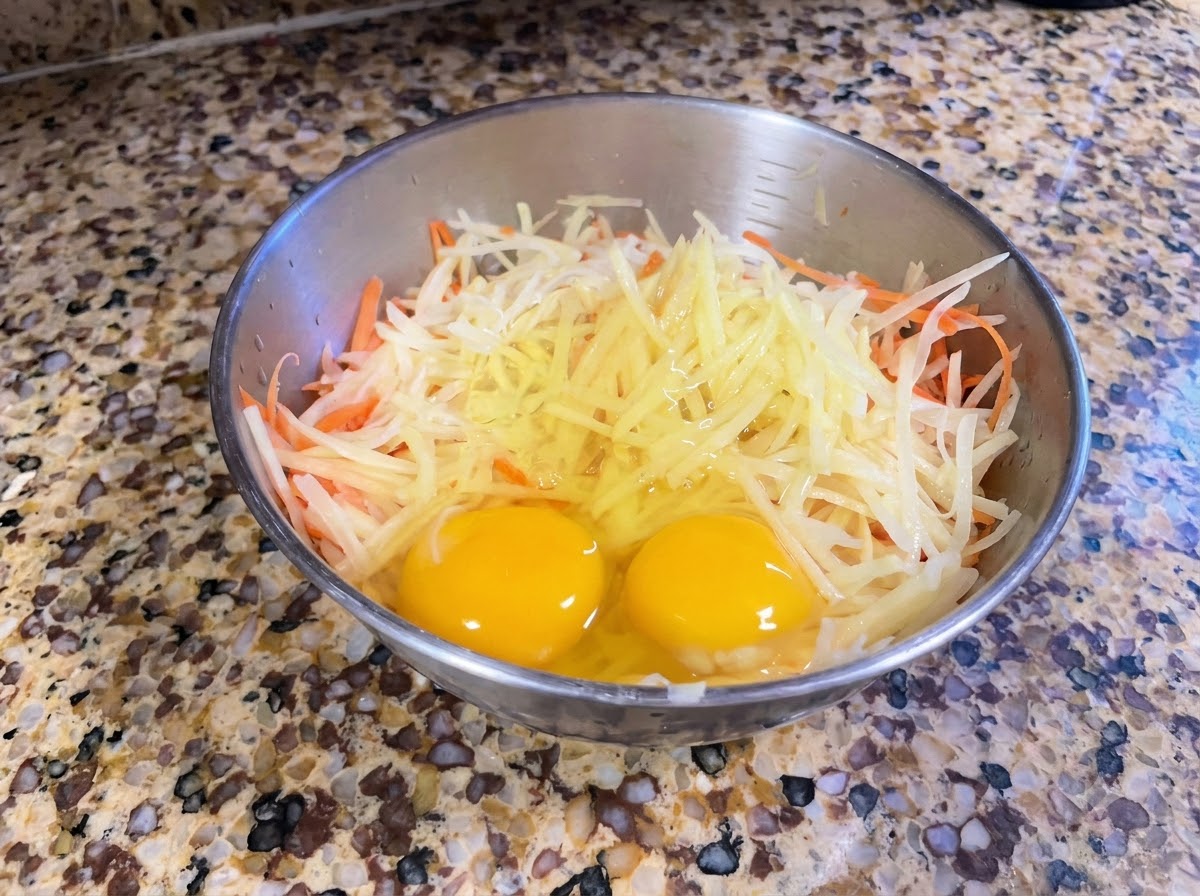 A metal bowl containing freshly grated potatoes and carrots with two whole eggs on top.