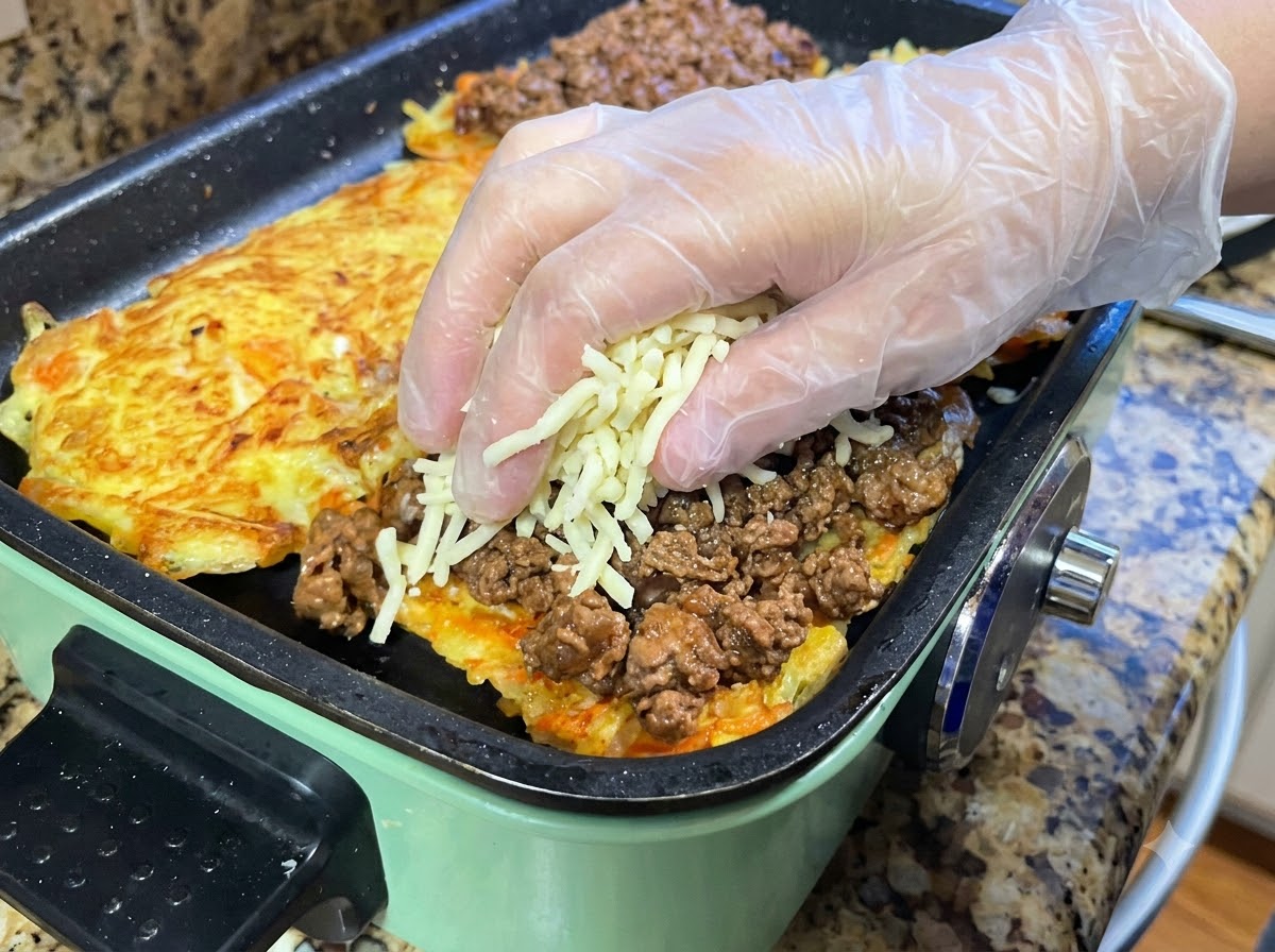 Hands wearing protective gloves placing a handful of shredded mozzarella cheese over a layer of cooked meat sauce spread across a pan-fried potato pancake.