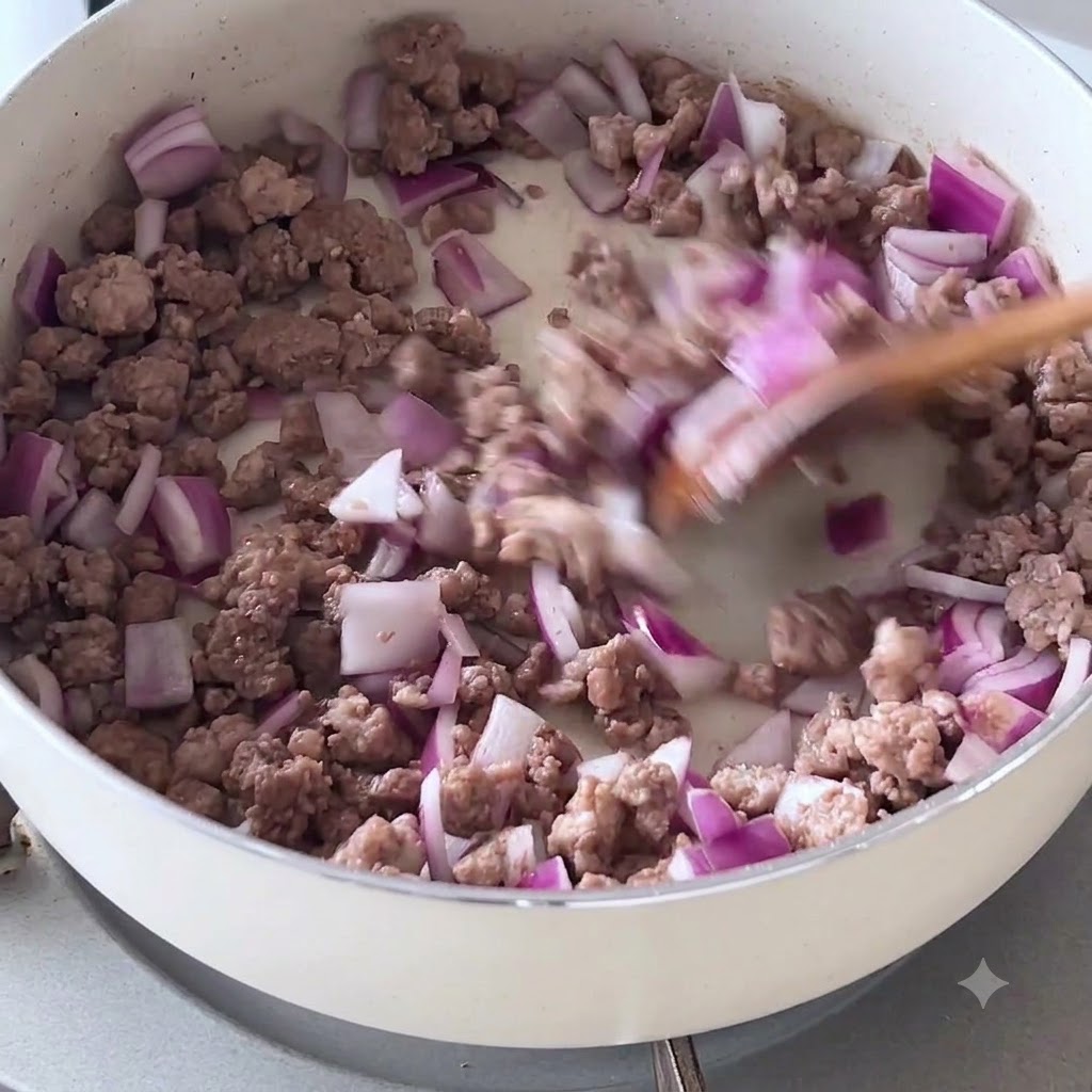 Chopped purple onions and browned minced beef being stirred with a wooden spatula in a white pan.