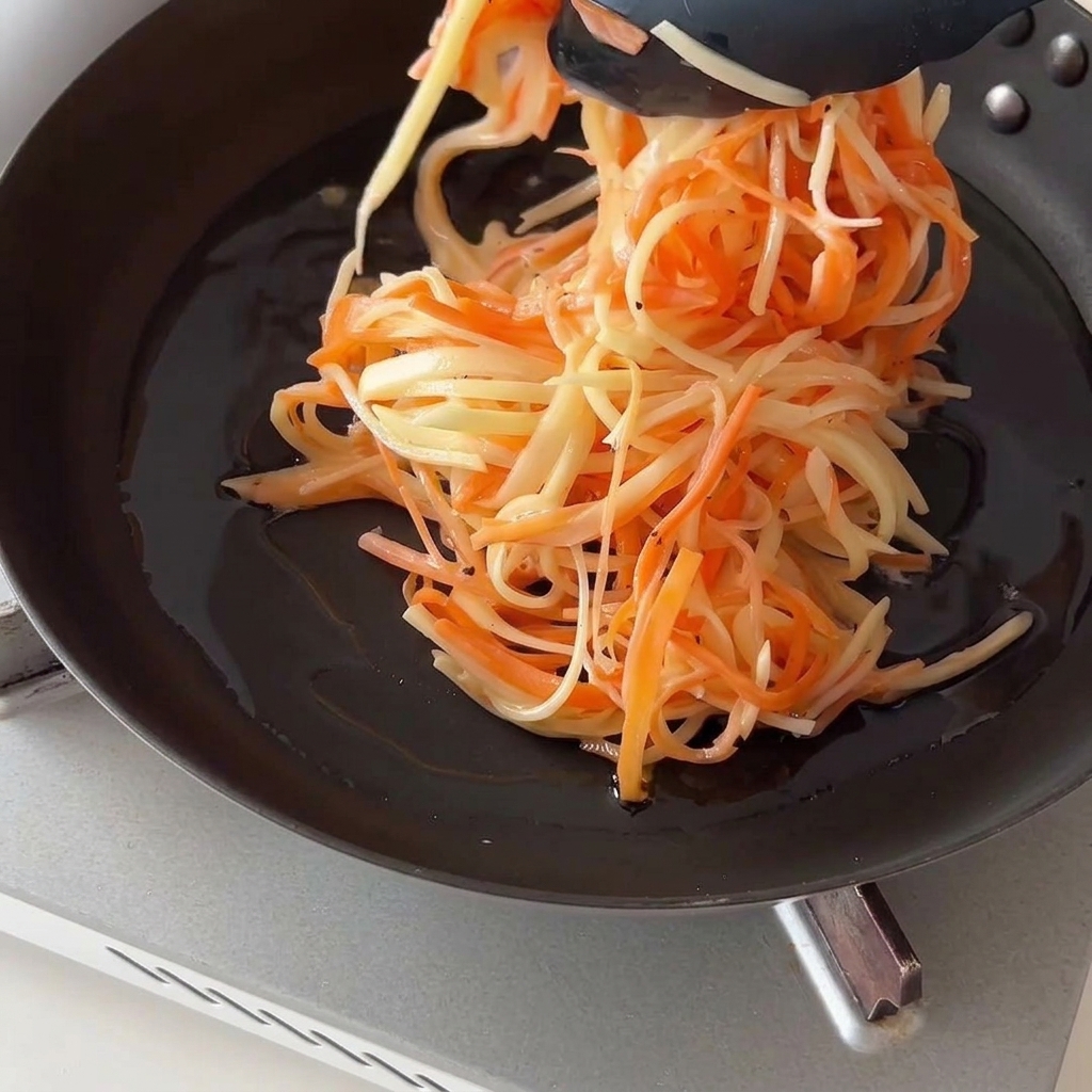 Using black kitchen tongs to place a mound of shredded potatoes and carrots into an oiled frying pan.