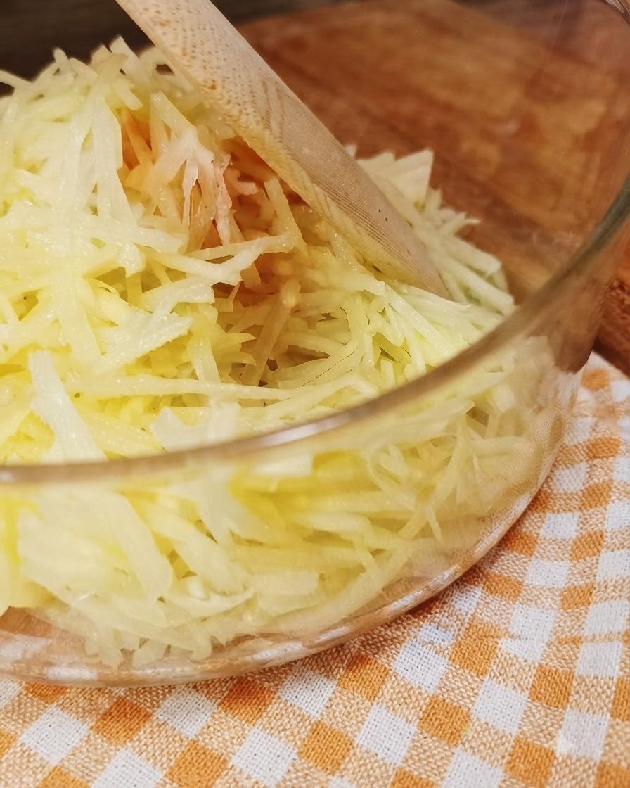 Freshly shredded potatoes inside a clear glass bowl with a wooden utensil.