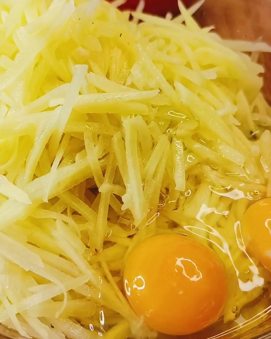 Whole eggs cracked directly over a bowl of raw shredded potatoes and seasonings.