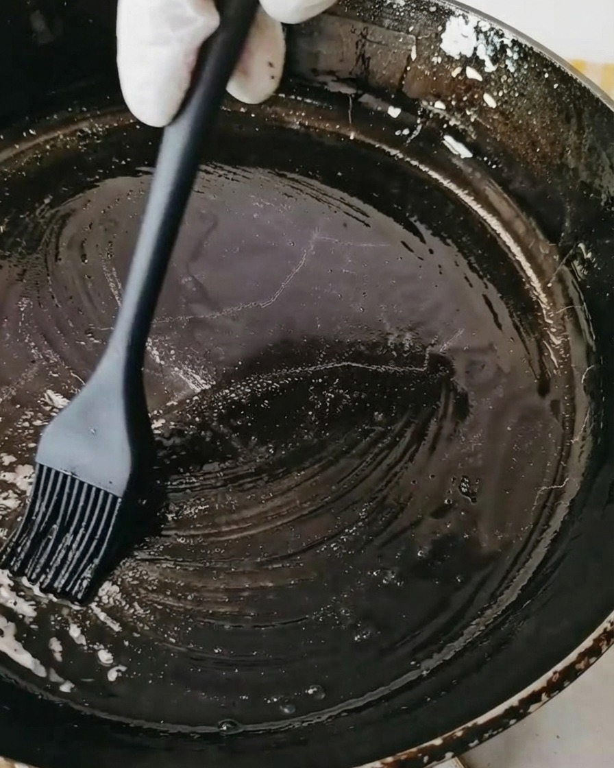 A black skillet being brushed with oil using a black silicone pastry brush.