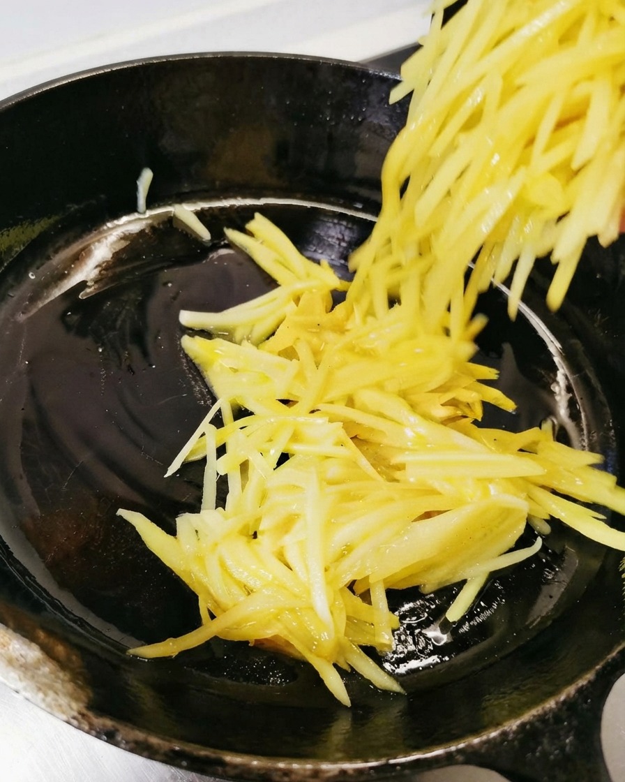 Shredded potatoes being poured and spread into the oiled black skillet.