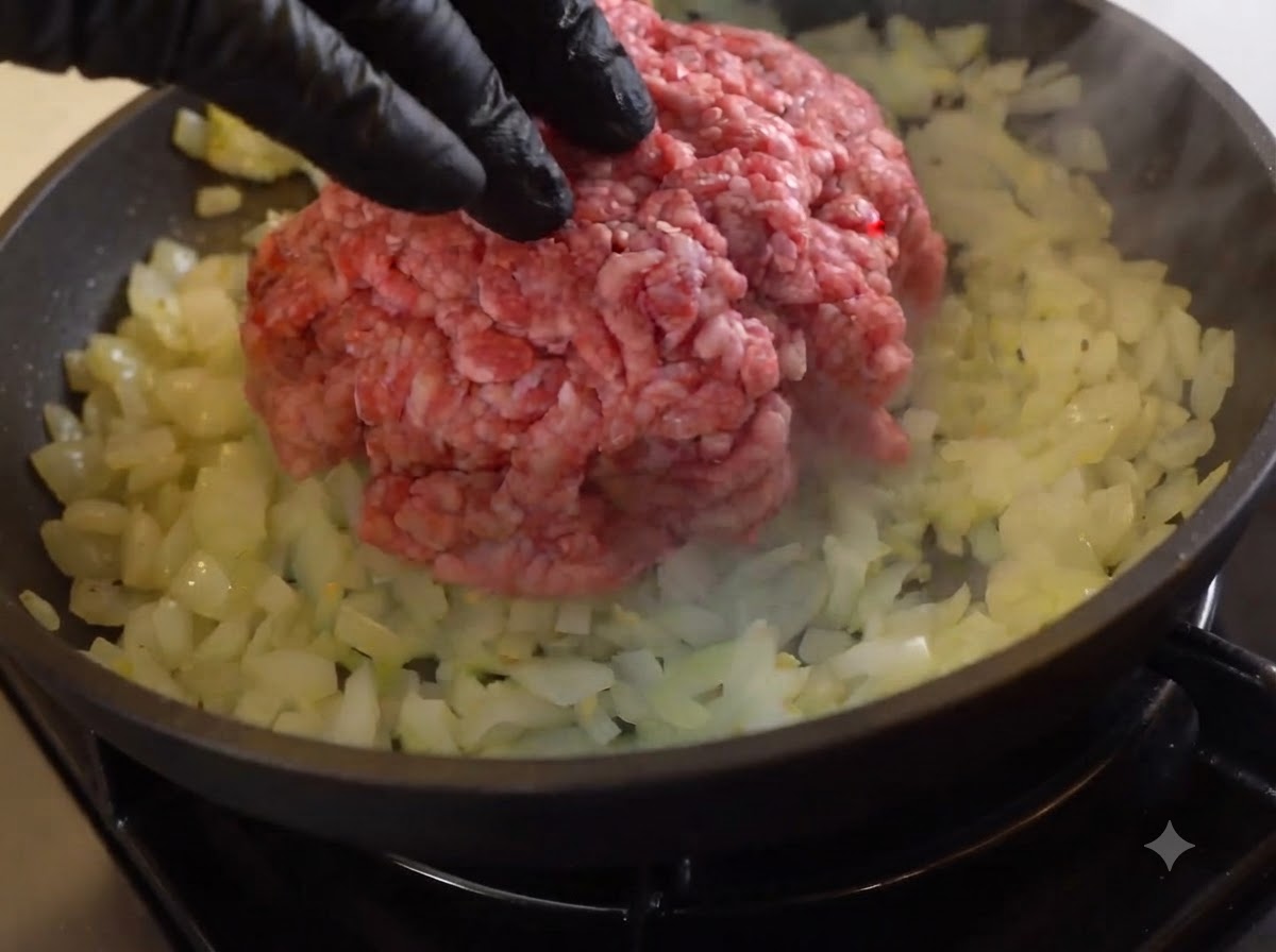 A mound of raw ground beef placed over sautéed onions in a skillet.