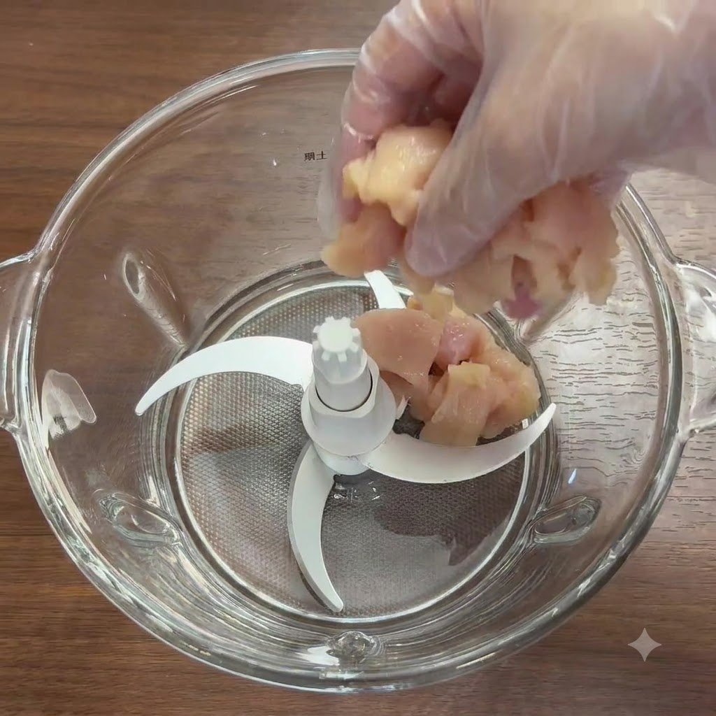 Chicken breast chunks and fresh vegetables inside a glass blender bowl with blades.