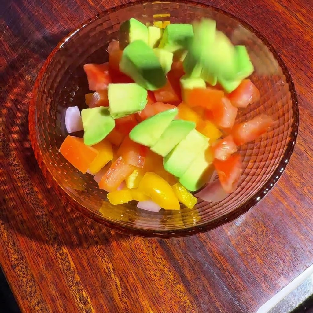 Diced vegetables and avocado in a glass bowl being tossed together with dressing.