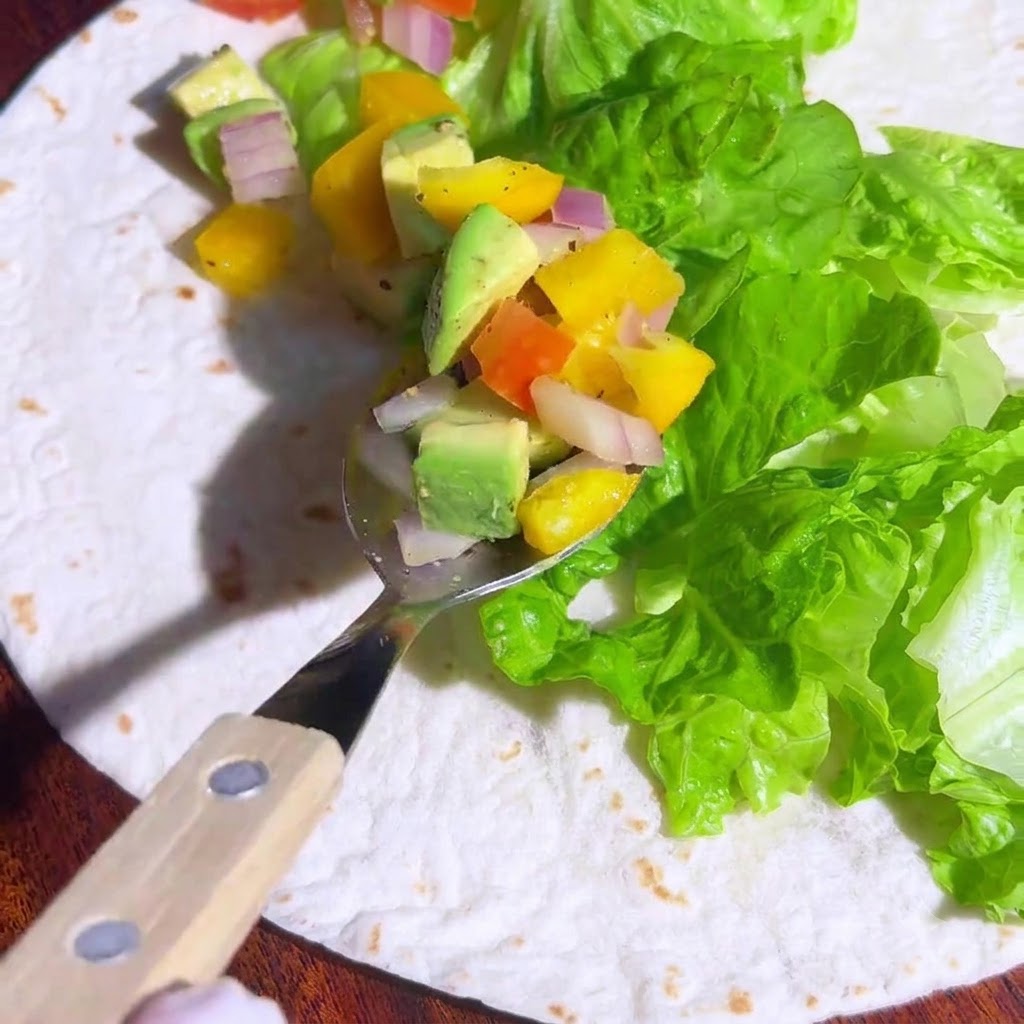 A spoon holding colorful diced avocado and vegetable salsa, positioned above the lettuce on a tortilla.