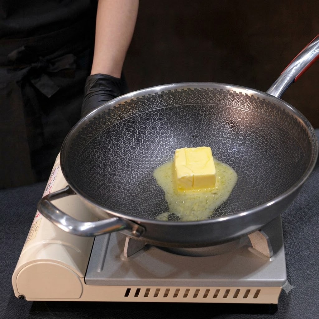 A block of yellow butter melting down in a metal wok, stirred with a small whisk over heat.
