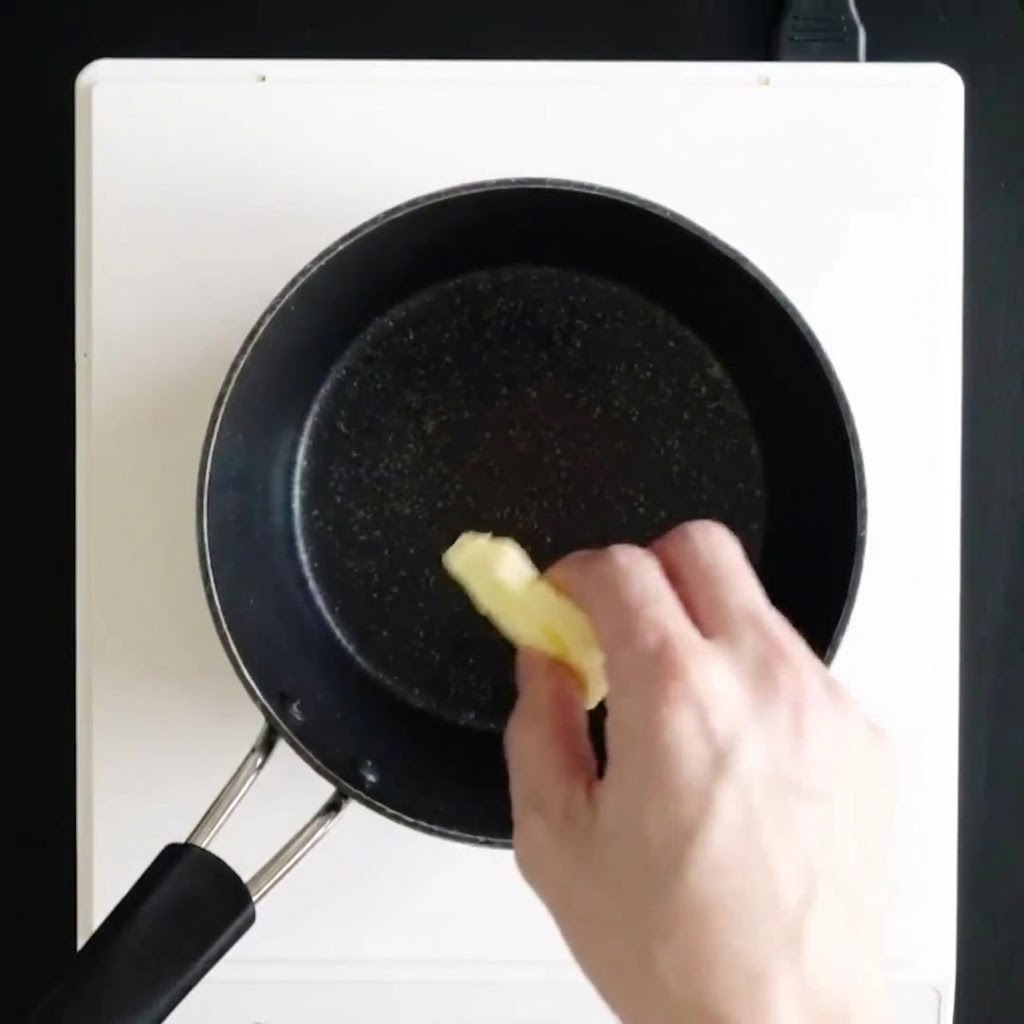A hand dropping pieces of butter into a black saucepan on a white stovetop to melt.