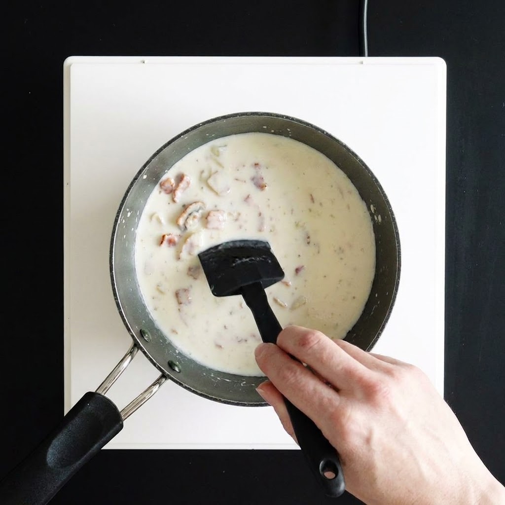 A hand stirring a thick, creamy white soup in a saucepan using a black silicone spatula.