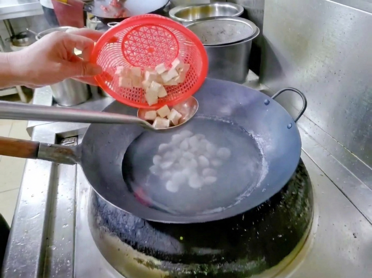 A hand holding a red strainer basket, dropping diced tofu into a wok of water and clams.