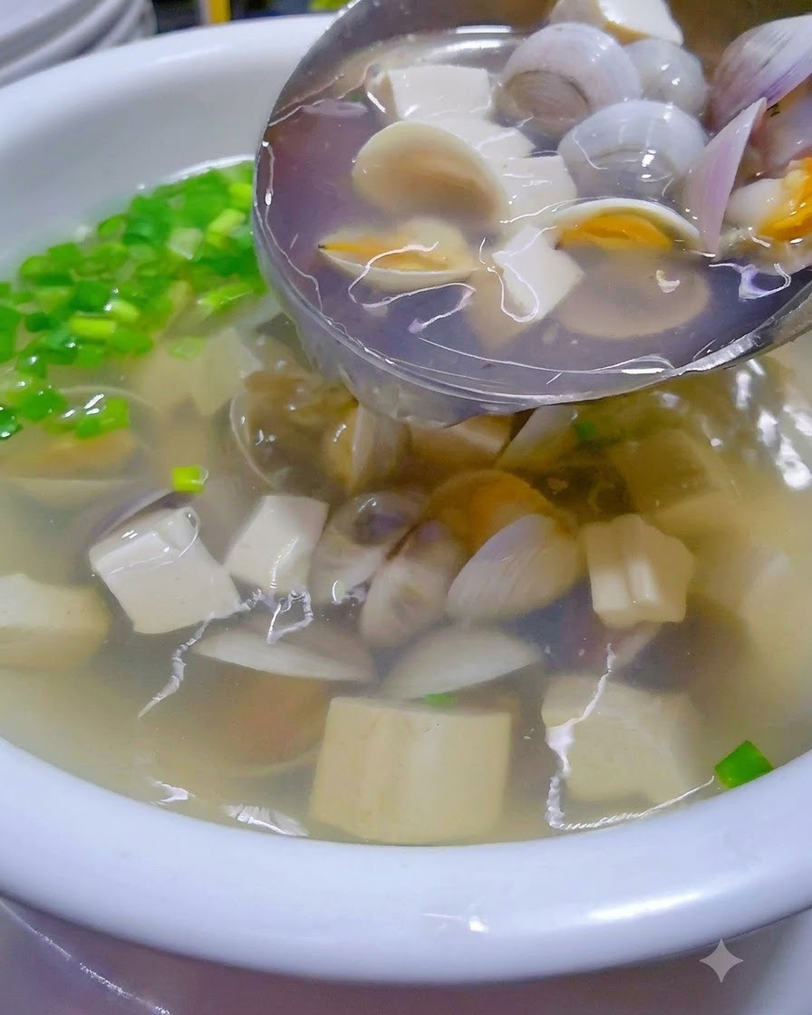 Hot clam and tofu soup being poured from a wok into a garnished serving bowl.