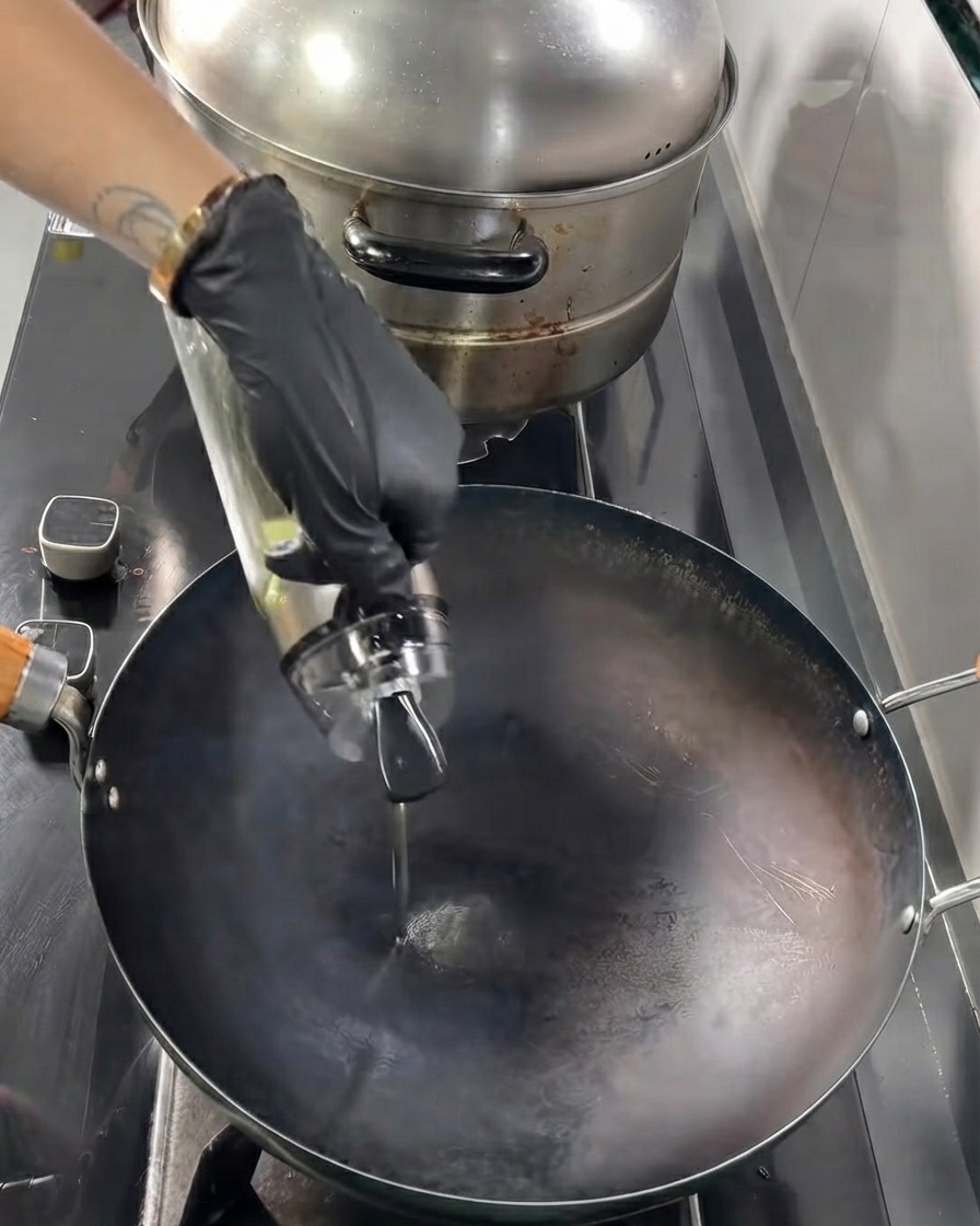 Cooking oil being poured from a glass bottle into a hot black wok.