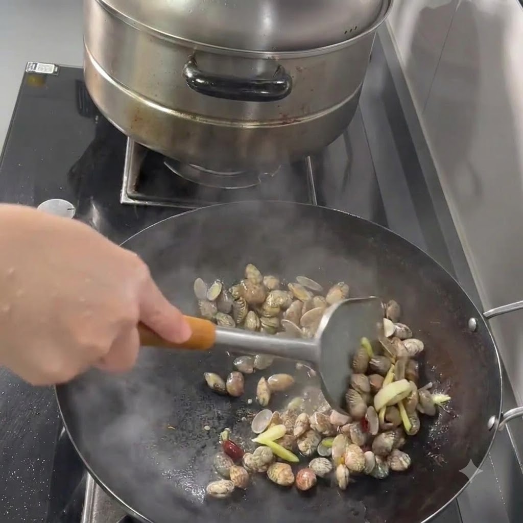 Clams, garlic, ginger, and chilies being stir-fried in a wok with a metal spatula.