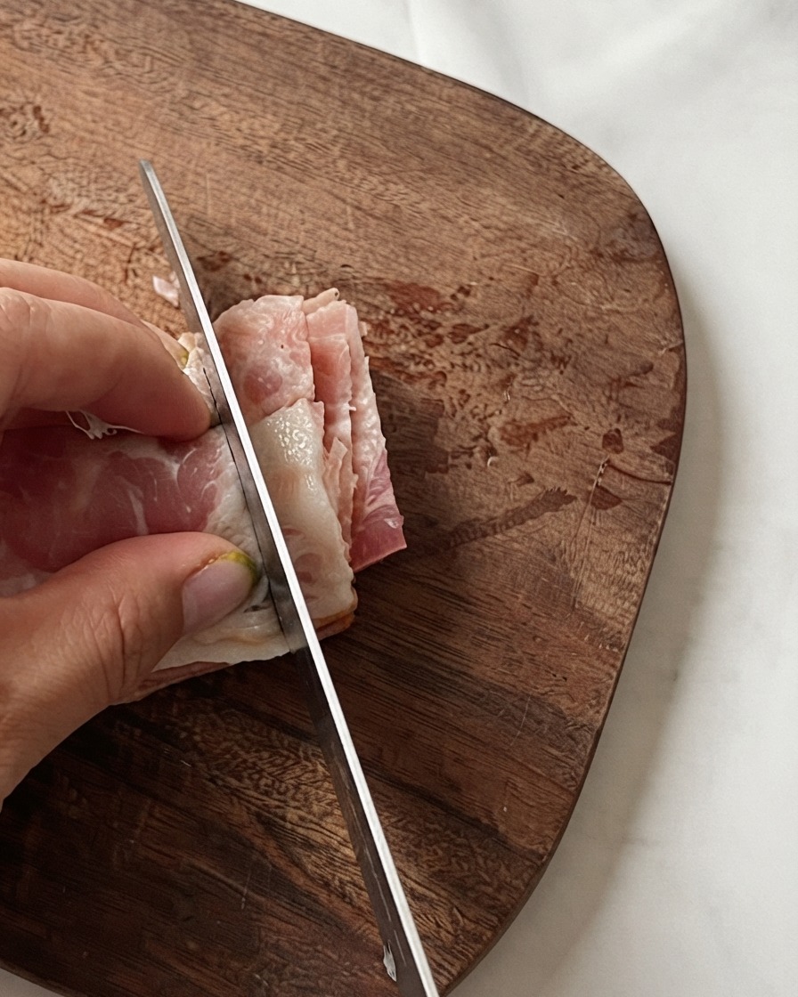 A chefs hand holding stacked bacon strips while a knife slices them into small segments on a wooden board.