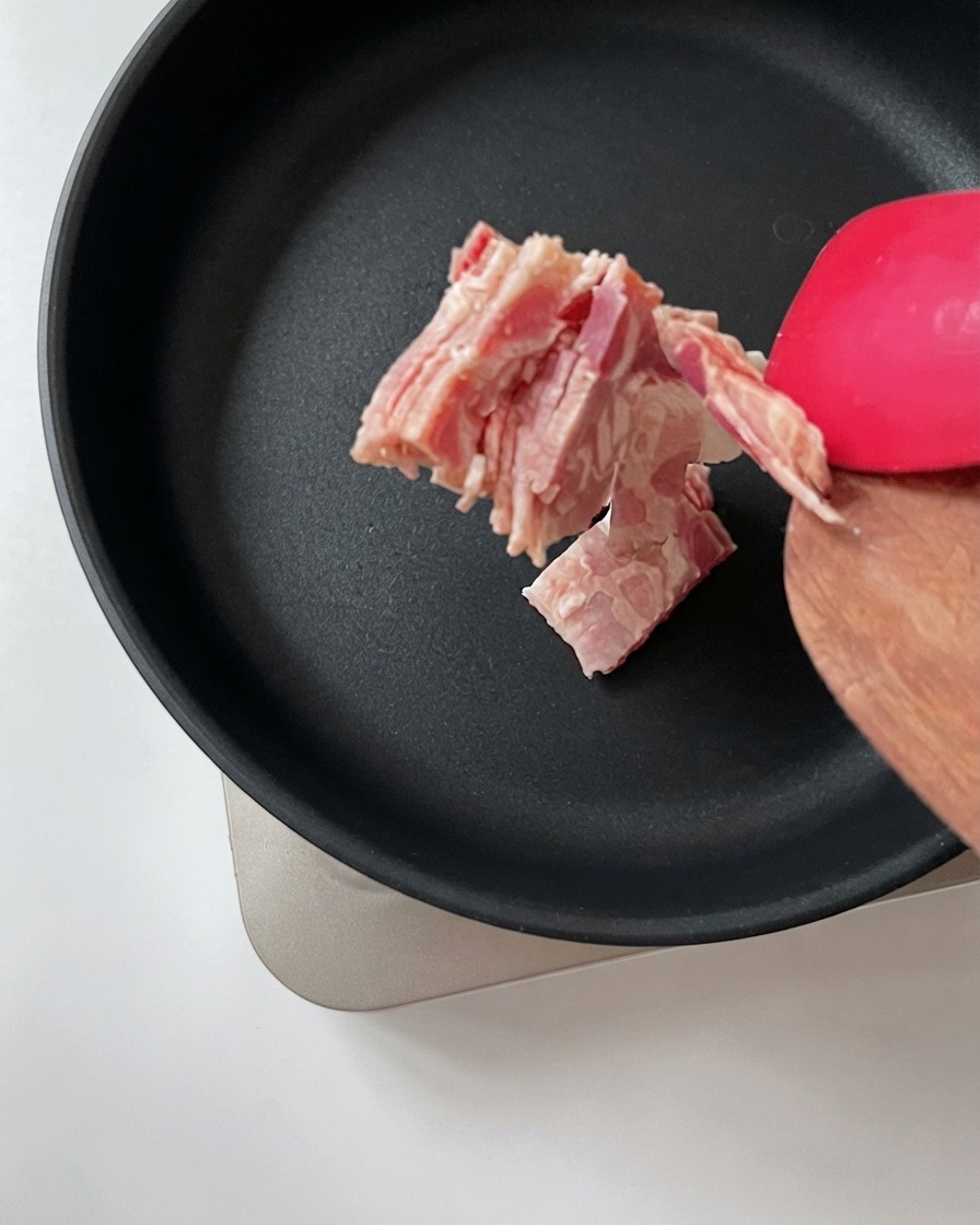 Chopped bacon being transferred from a wooden spatula into a hot black frying pan.