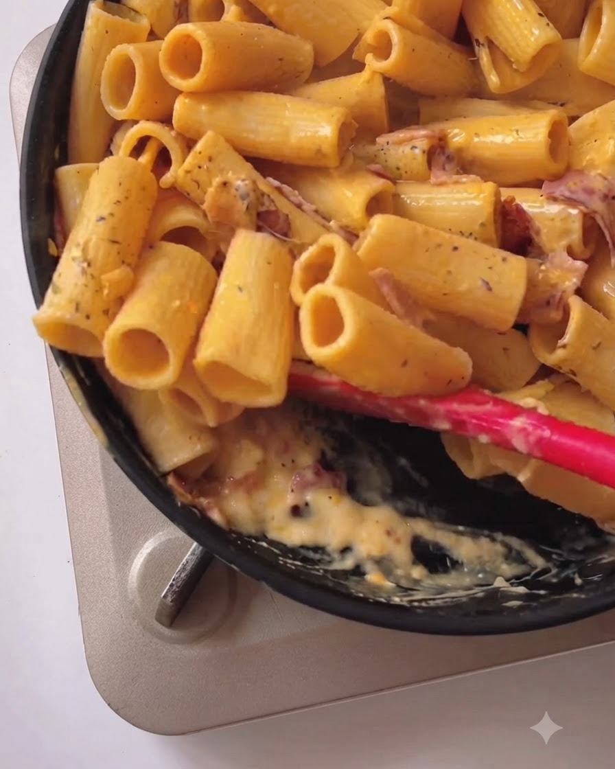 Rigatoni pasta being tossed and mixed with creamy yellow sauce and bacon in a black pan using a red silicone spatula.