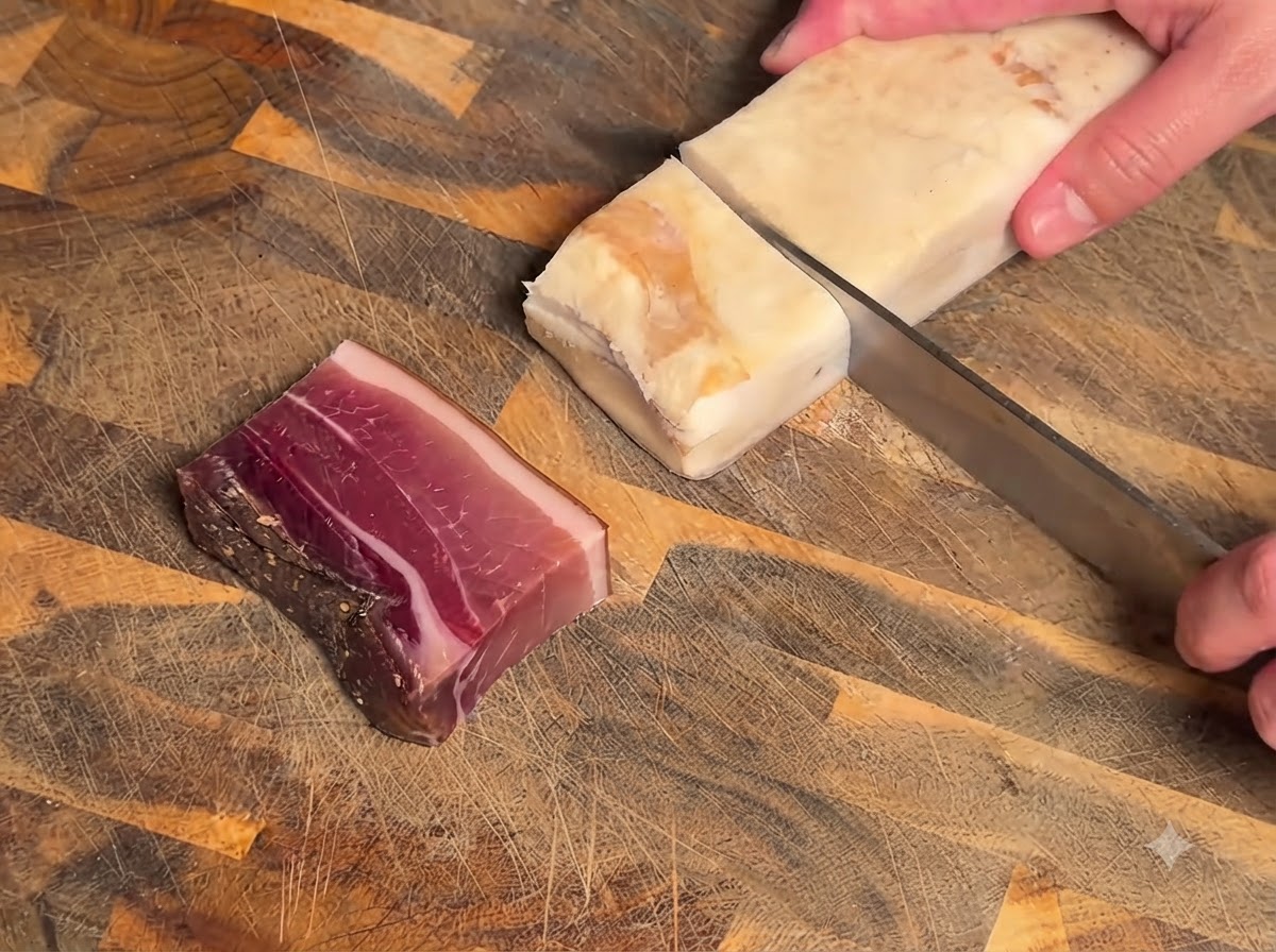 A knife slicing a block of cured meat and bacon on a textured wooden cutting board.