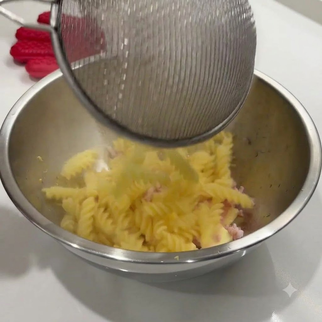 Cooked spiral pasta being poured from a wire strainer into a stainless steel bowl with ingredients.