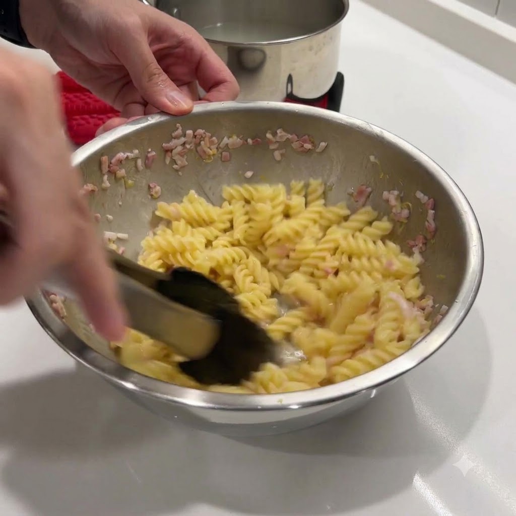 Stirring hot spiral pasta and butter mixture in a metal bowl using black tongs.