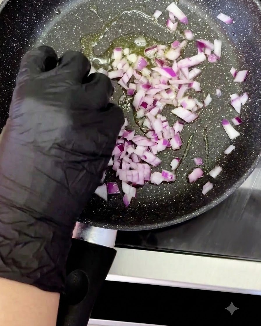 A gloved hand adding diced red onion into a heated pan with sizzling oil.
