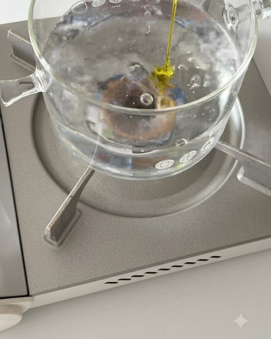 Rigatoni pasta boiling in a glass pot on a stove with a stream of olive oil being poured in.