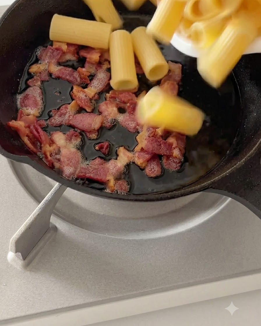 Boiled rigatoni pasta being tossed with cooked bacon in a skillet over low heat.