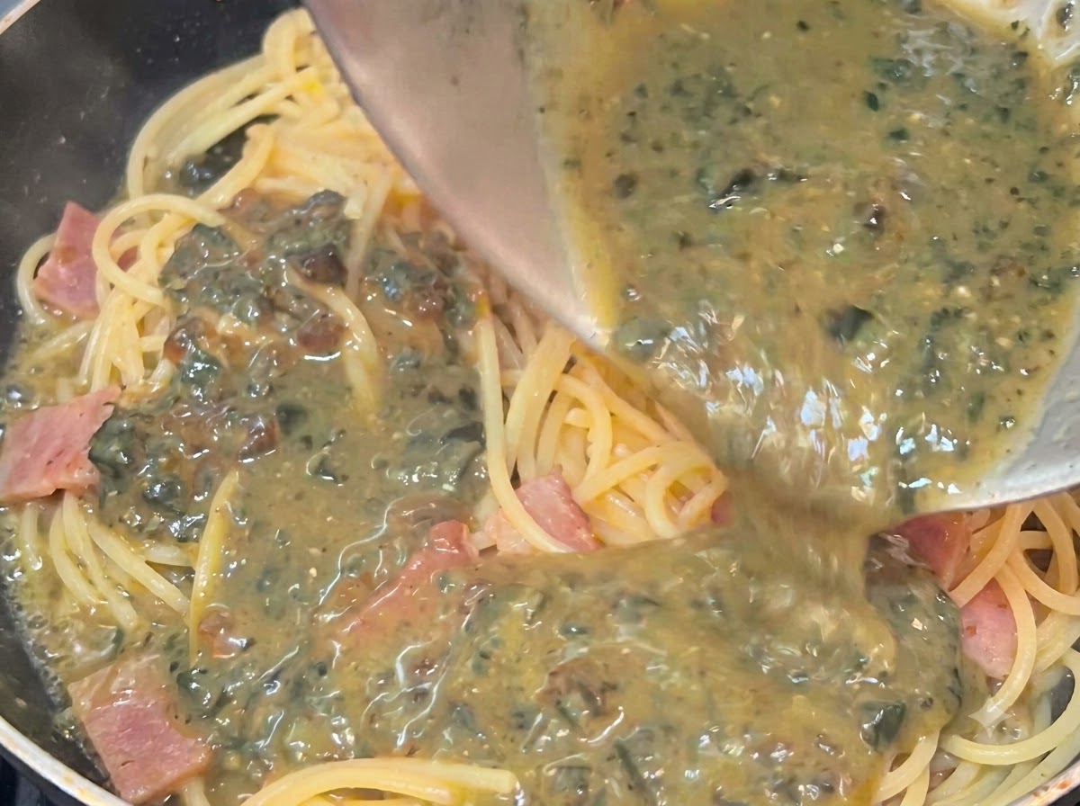 Creamy, speckled preserved egg sauce being poured from a bowl over hot spaghetti and bacon in a pan.