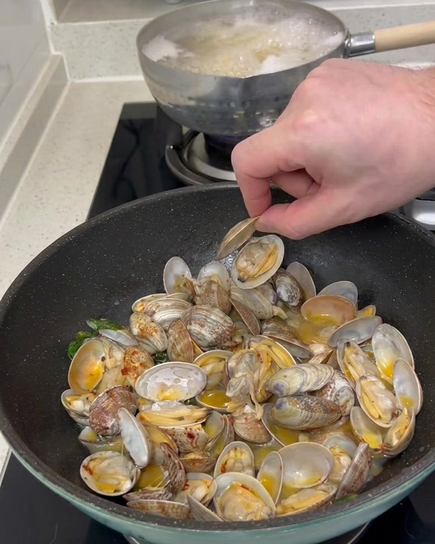 A persons hand picking up an open clam shell from a pan filled with cooked clams and sauce.
