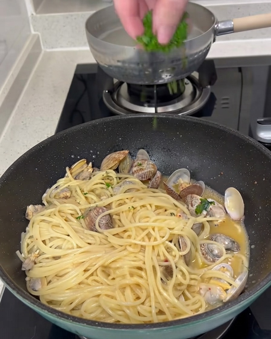Fresh chopped green parsley being sprinkled from above onto a pan of cooked spaghetti and clams.
