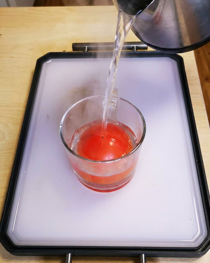 Hot water being poured from a kettle into a glass cup containing a fresh red tomato.