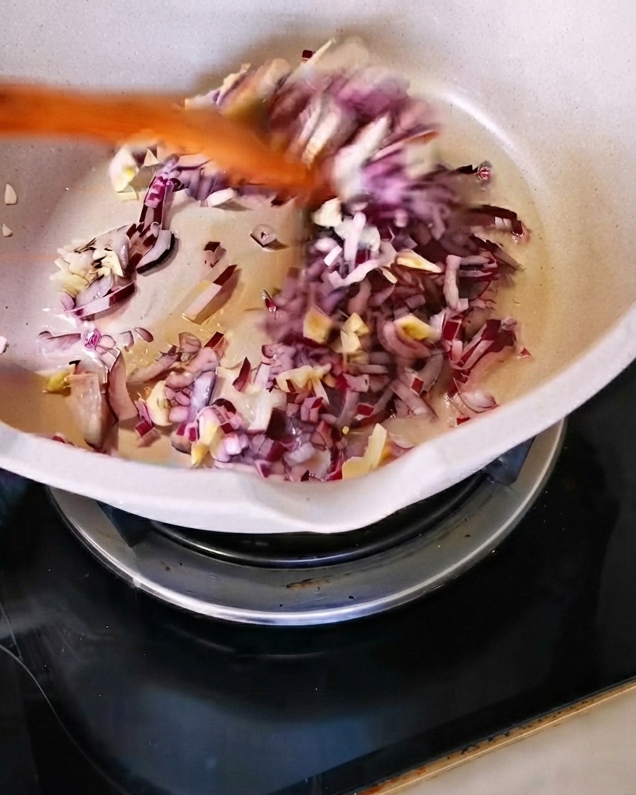 Chopped red onion and minced garlic being sautéed with a wooden spatula in a white pan.