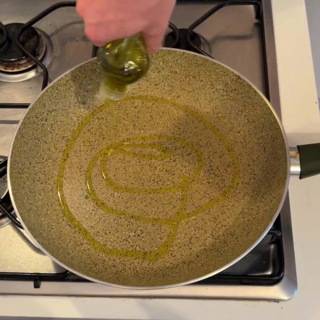 A person pouring olive oil from a bottle into a large speckled pan on a gas stovetop.