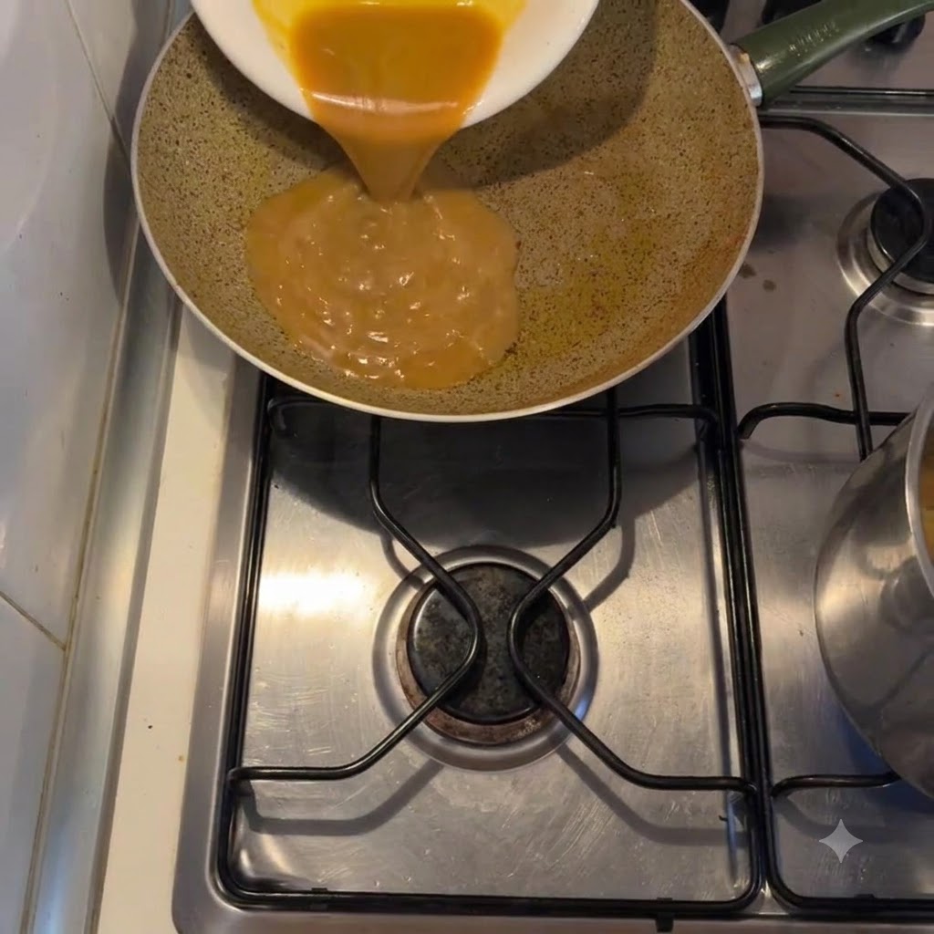 Thick golden sauce being poured from a white plate into a frying pan on a stovetop.