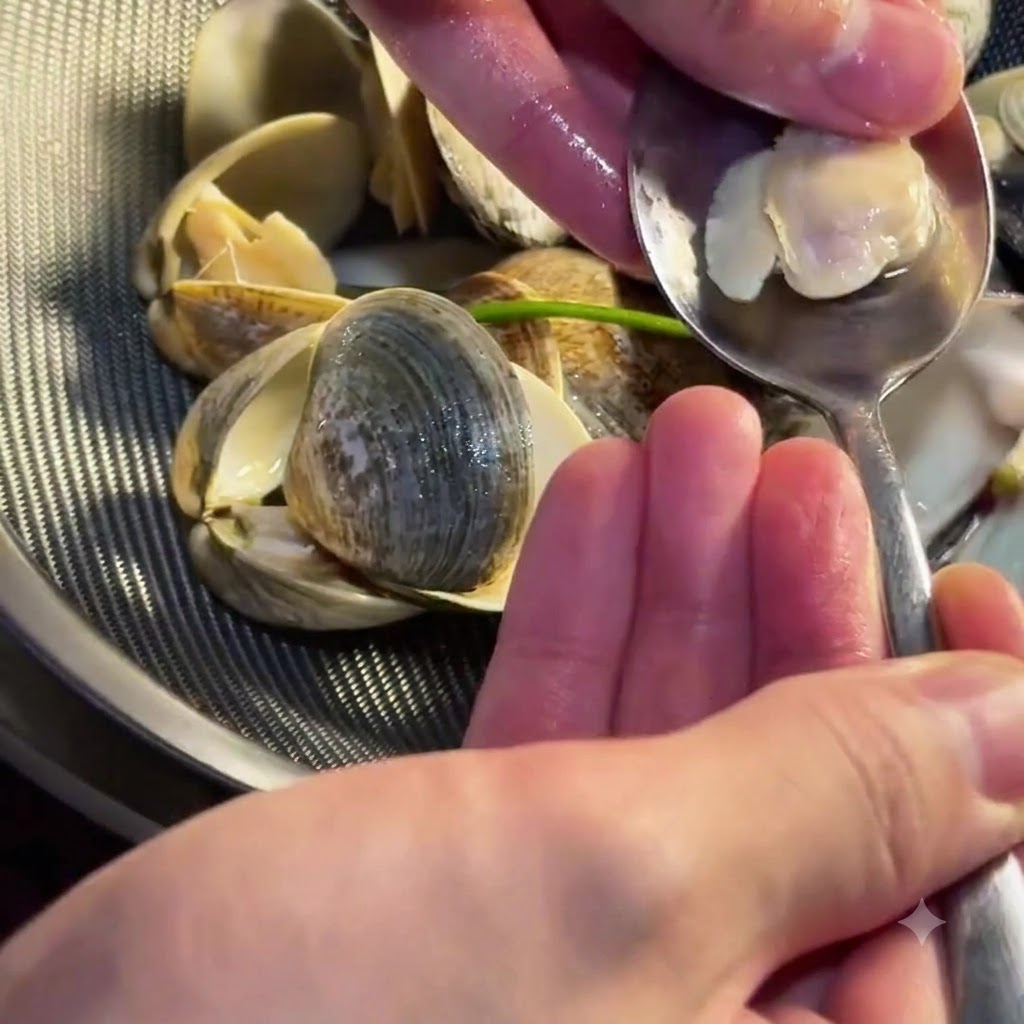 A person using a small spoon to scoop tender clam meat out of its shell.