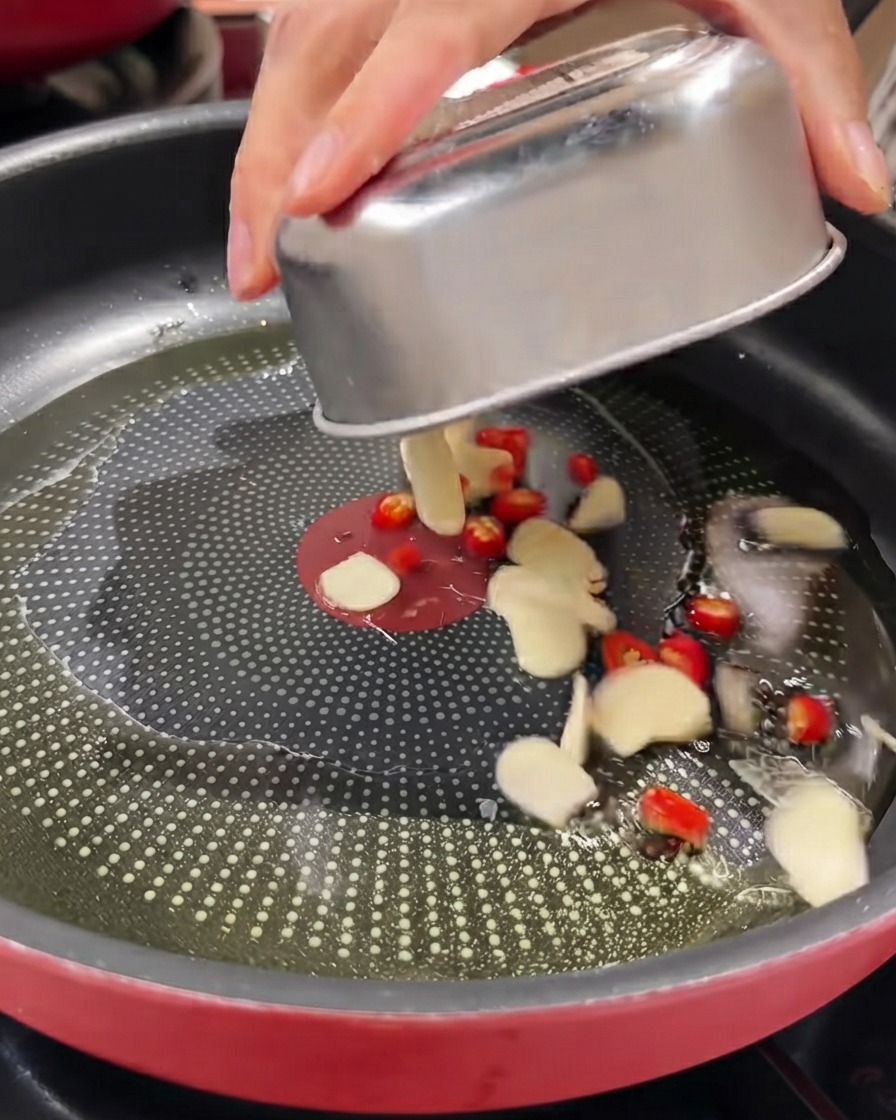 Sliced garlic, red birds eye chilies, and chives being tipped from a metal container into an oiled pan.