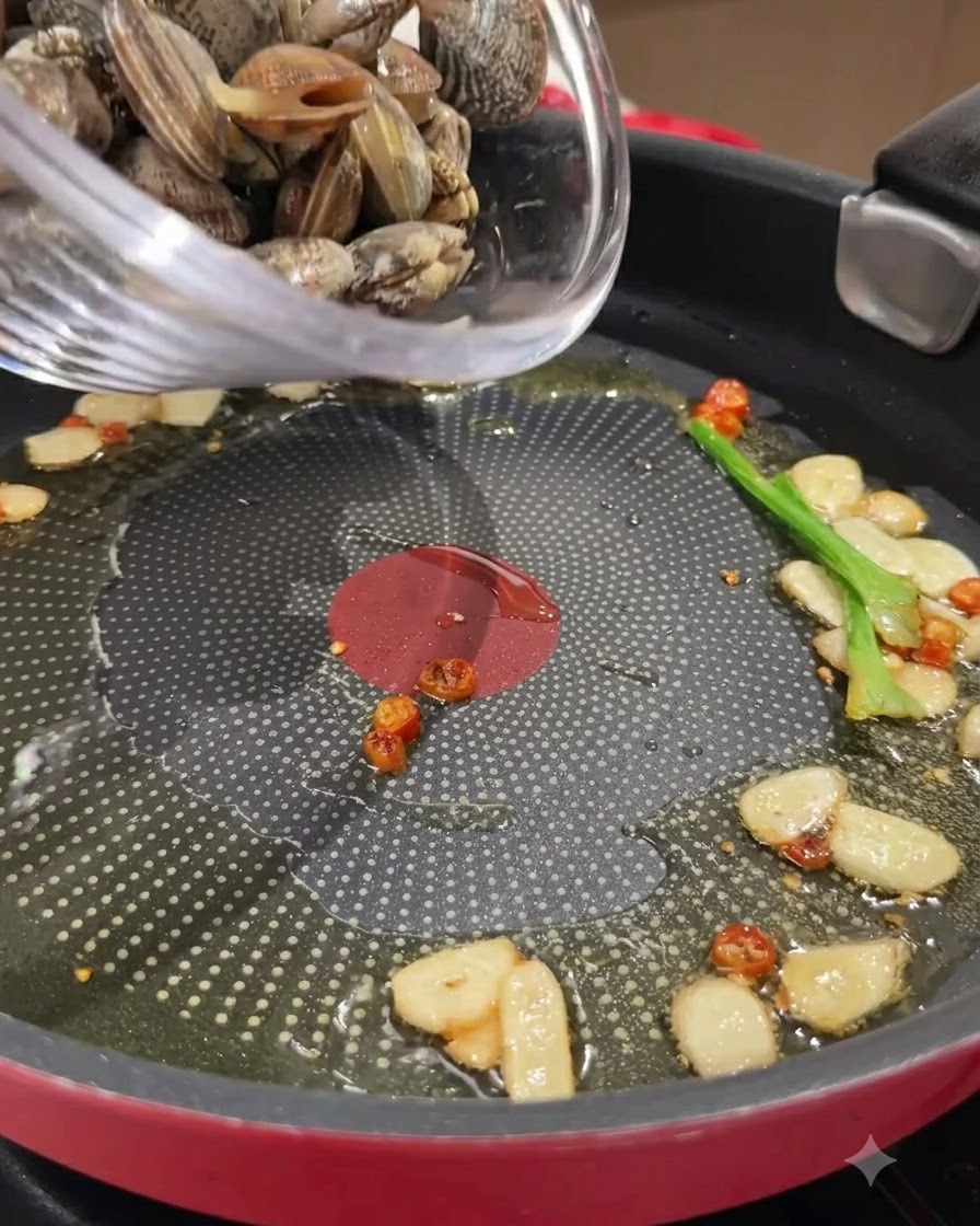 Fresh clams being poured from a glass bowl into a frying pan with sautéed garlic and chilies in oil.