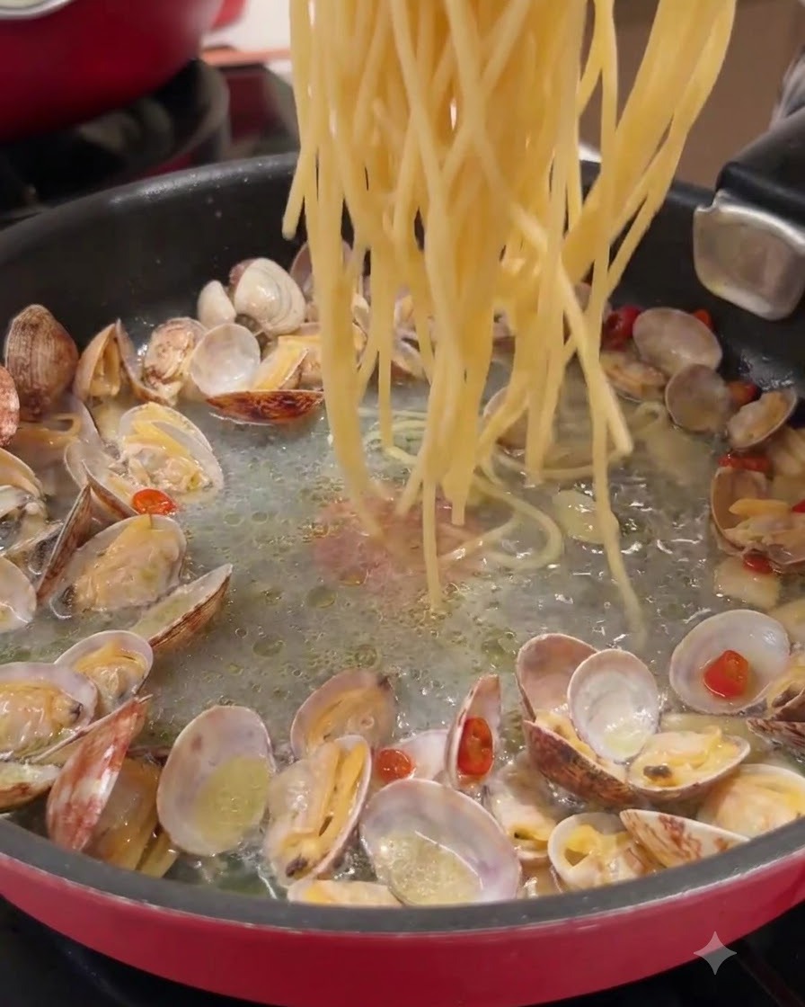 Long strands of pasta being dropped into a pan containing cooked clams and seasoned liquid.