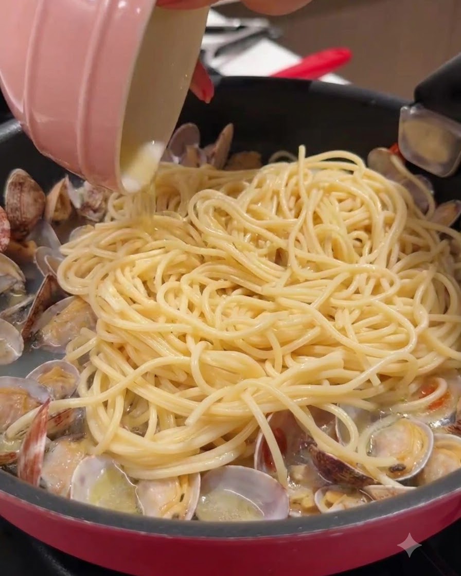 A small pink bowl pouring liquid into a pan filled with spaghetti and clams.