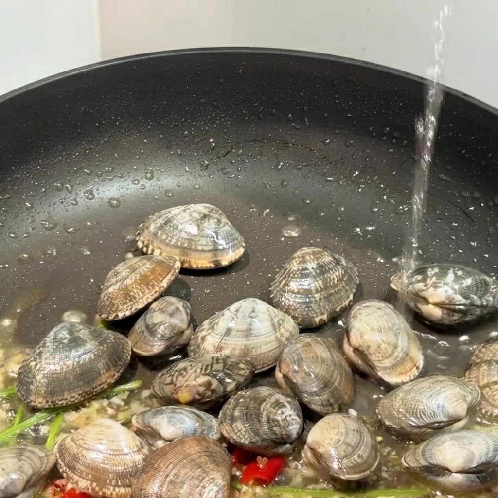 A black skillet containing clams, garlic, and chili peppers with liquid being poured into it.