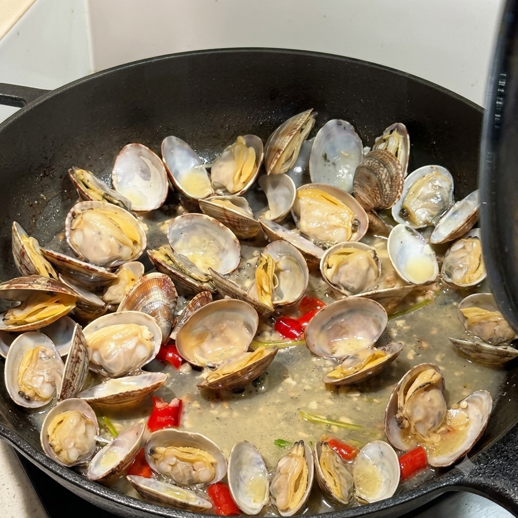 Opened clams cooking in broth inside a skillet with a lid partially visible.