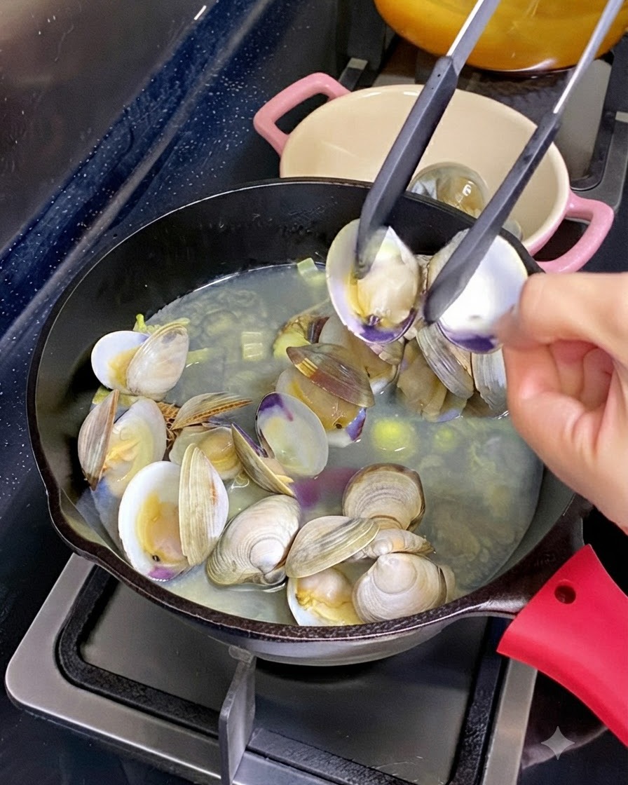 A pair of kitchen tongs holding open a clam shell with cooked meat inside, above a skillet of simmering clams and broth.